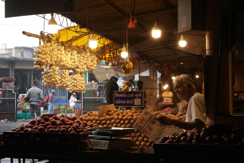 A vendor reads a newspaper in a bustling Indian market filled with fresh produce and vibrant ambiance.